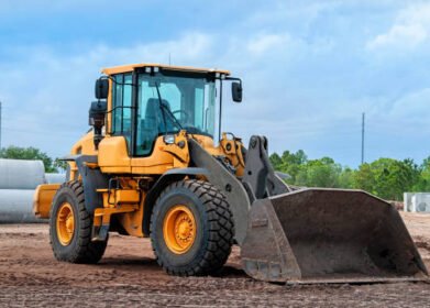 construction site with wheel loader in foreground, sewer pipe and tree line in background, sunny day, no people, machinery on dirt with bucket down on ground, blue sky with some clouds
